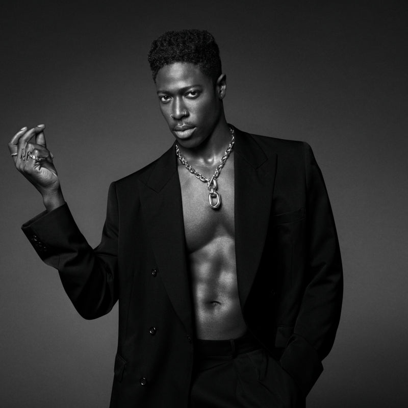 Man wearing a black suit with a chain necklace against a dark background