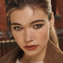 Close-up of a woman with a neutral expression, wearing a brown coat.