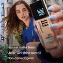 Woman holding a Maybelline Fit Me foundation bottle with product features displayed.