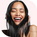 A close-up portrait of a smiling woman with long, straight black hair, closed eyes, and a joyful expression against a pale pink background.