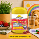 Nature's Way Kids Smart Vita Gummies Iron + Vitamin C supplement bottle displayed on a wooden table with strawberries, blueberries, a glass of water, and children's room decor in the background.