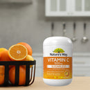 A bottle of Nature's Way Vitamin C 500mg sugarless supplement stands on a kitchen counter next to a black basket filled with oranges, with a blurred kitchen background.