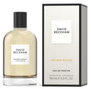 A bottle of David Beckham Refined Woods Eau de Parfum with the cap facing forward, next to its packaging box. The bottle is beige and the box is black and white with the brand's logo.