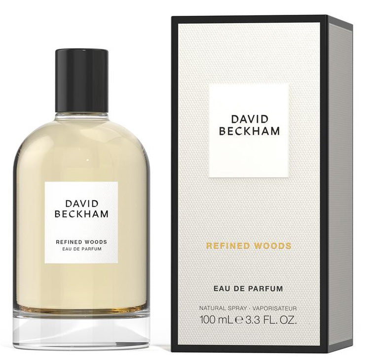 A bottle of David Beckham Refined Woods Eau de Parfum with the cap facing forward, next to its packaging box. The bottle is beige and the box is black and white with the brand's logo.
